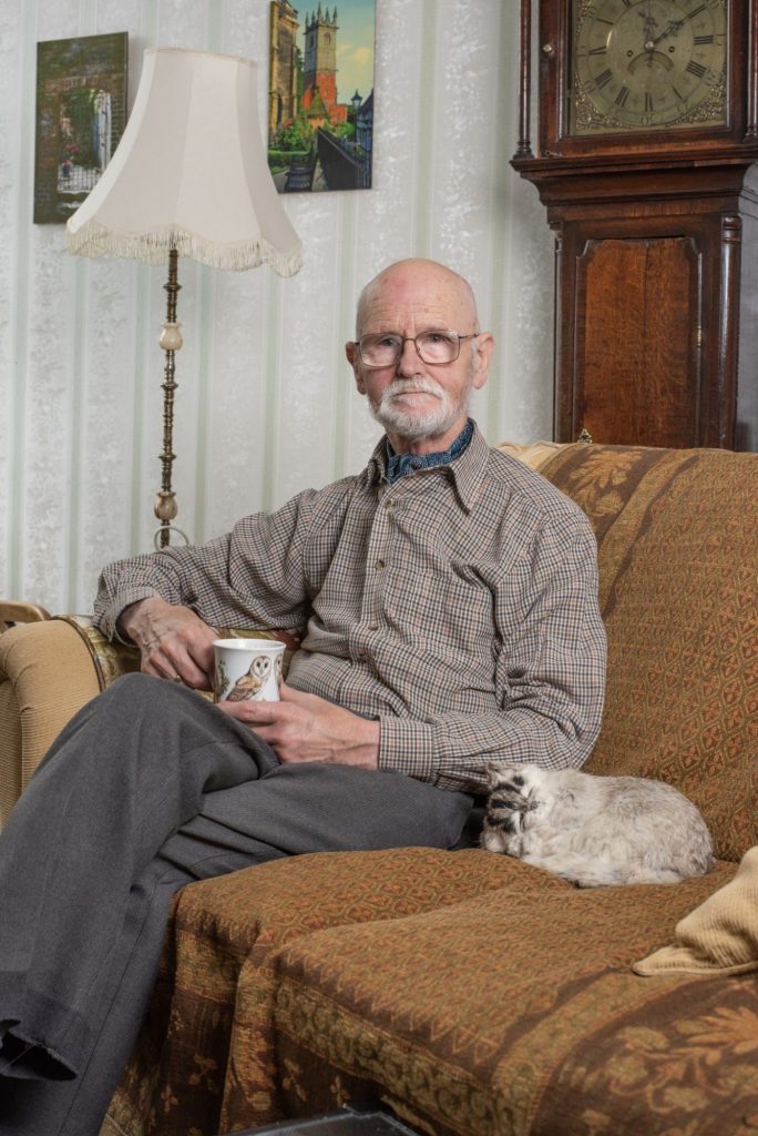 Portrait of an older man with a white beard and glasses, sitting on a patterned sofa with a mug, beside a grandfather clock