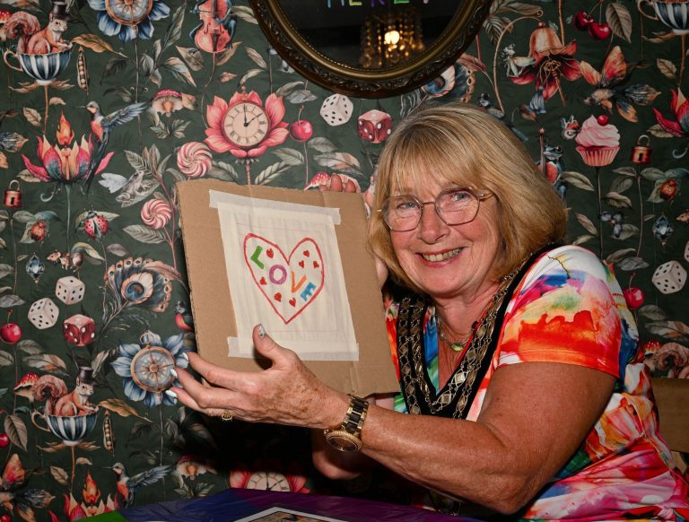 A smiling woman shows off a heart she has stitched at a craft session in Cheadle