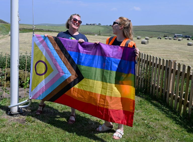 Two volunteers hold up the Progress Pride flag in a field at Flash