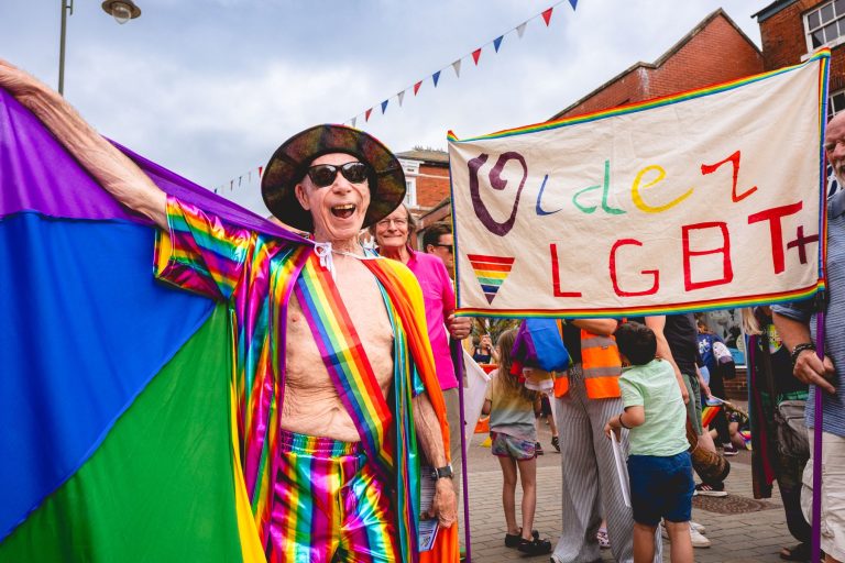 A marcher in a rainbow cape beside the Older LGBT+ banner in Leek