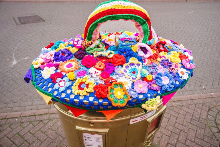 A yarn-bombed post box topper covered in crocheted flowers and a rainbow in Leek