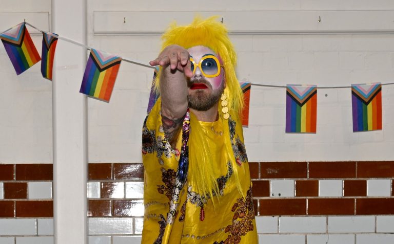 A drag performer in a yellow wig and sunglasses poses beside Pride bunting