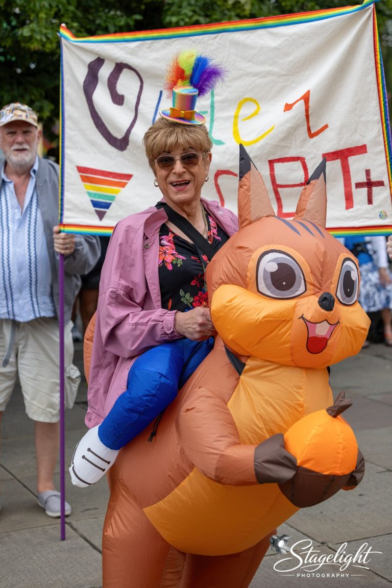 A marcher appears to ride an inflatable orange squirrel beside the Older LGBT+ banner in Leek