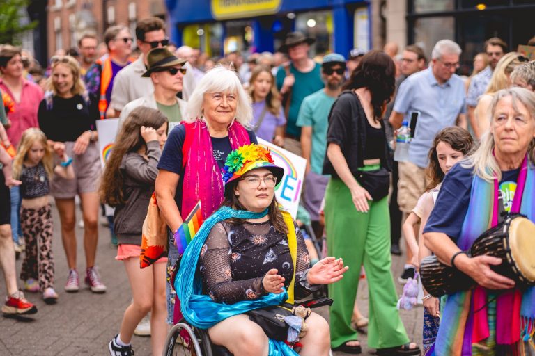Marchers, including a smiling wheelchair user in a rainbow hat, join the Pride parade through Leek